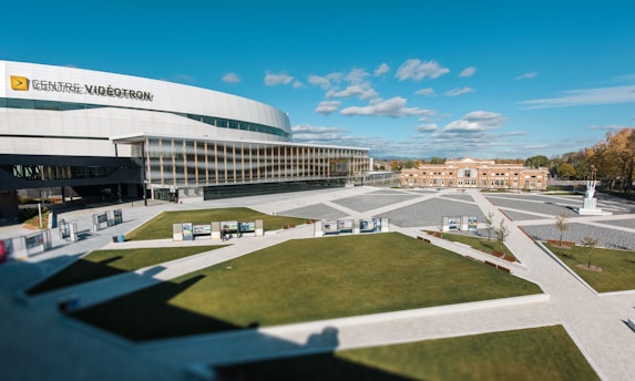 A large, modern arena bearing the sign 'Centre Vidéotron' with a sleek, curved architectural design. The building is surrounded by an open plaza featuring geometric patterns in the pavement and well-maintained grassy areas. A smaller, brick building is visible in the background against a bright blue sky with scattered clouds.