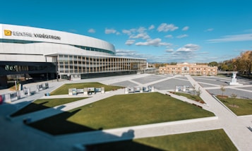 A large, modern arena bearing the sign 'Centre Vid&eacute;otron' with a sleek, curved architectural design. The building is surrounded by an open plaza featuring geometric patterns in the pavement and well-maintained grassy areas. A smaller, brick building is visible in the background against a bright blue sky with scattered clouds.