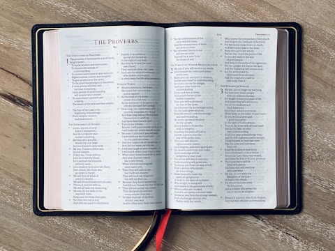An open book resting on a wooden surface, displaying text from Proverbs. The pages are neatly aligned, with a red bookmark visible coming out from the bottom. The book's left and right pages have columns of text.