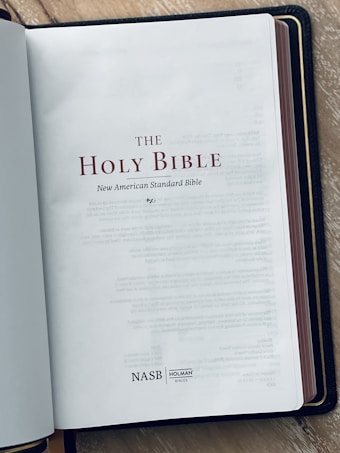 The image displays an open book with the title page reading 'THE HOLY BIBLE' and 'New American Standard Bible'. There is a logo 'NASB' and another for 'HOLMAN BIBLES' at the bottom of the page. The background surface appears to be a wooden texture.
