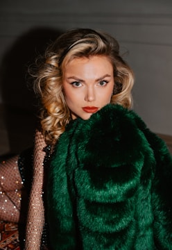 A person with curly blonde hair and bold makeup gazes directly at the camera. The individual is draped in a luxurious green fur coat. They are seated indoors, and part of their outfit includes sparkly mesh sleeves.