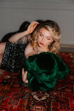 A woman with styled blonde hair is lying on a richly patterned Persian carpet. She is wearing a black, mesh-like dress embellished with sequins and holding a luxurious green fur or fabric. Her makeup is elegantly done with red lipstick and eyeliner.