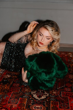 A woman with styled blonde hair is lying on a richly patterned Persian carpet. She is wearing a black, mesh-like dress embellished with sequins and holding a luxurious green fur or fabric. Her makeup is elegantly done with red lipstick and eyeliner.
