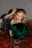 A woman with styled blonde hair is lying on a richly patterned Persian carpet. She is wearing a black, mesh-like dress embellished with sequins and holding a luxurious green fur or fabric. Her makeup is elegantly done with red lipstick and eyeliner.