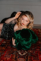 A woman with styled blonde hair is lying on a richly patterned Persian carpet. She is wearing a black, mesh-like dress embellished with sequins and holding a luxurious green fur or fabric. Her makeup is elegantly done with red lipstick and eyeliner.