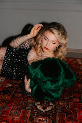 A woman with styled blonde hair is lying on a richly patterned Persian carpet. She is wearing a black, mesh-like dress embellished with sequins and holding a luxurious green fur or fabric. Her makeup is elegantly done with red lipstick and eyeliner.