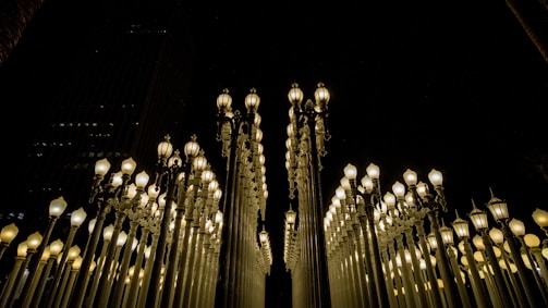 A multitude of vintage-style street lamps are arranged in a symmetrical pattern against a dark night sky. The lamps are lit, emitting a warm glow that contrasts with the surrounding darkness.
