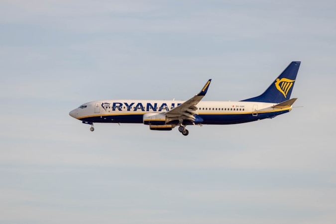A commercial airplane flies through a clear sky. The aircraft is painted in Ryanair's blue and white livery, with the company's logo prominently visible on the tail.