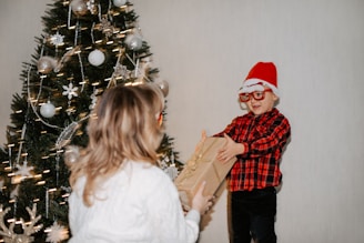 A child wearing a red Santa hat and glasses, dressed in a red and black plaid shirt, is handing a gift to another person whose back is facing the camera. The setting includes a decorated Christmas tree adorned with ornaments and lights.
