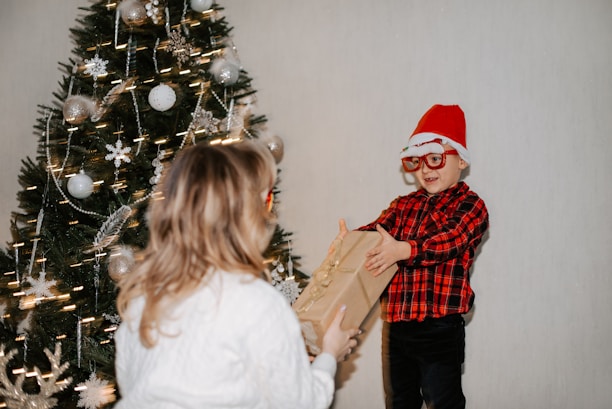 A child wearing a red Santa hat and glasses, dressed in a red and black plaid shirt, is handing a gift to another person whose back is facing the camera. The setting includes a decorated Christmas tree adorned with ornaments and lights.