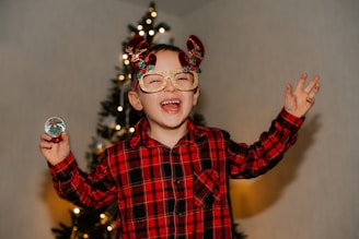 A toddler wearing a red and green Christmas sweater with reindeer patterns, smiling by a decorated Christmas tree.
