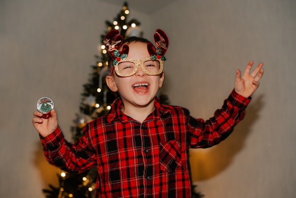 A toddler wearing a red and green Christmas sweater with reindeer patterns, smiling by a decorated Christmas tree.