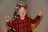 A child wearing a red and black plaid shirt and festive antler glasses stands in front of a decorated Christmas tree. The child is smiling widely and holding a small snow globe in one hand with an expression of joy.