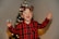 A child wearing a red and black plaid shirt and festive antler glasses stands in front of a decorated Christmas tree. The child is smiling widely and holding a small snow globe in one hand with an expression of joy.