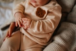A close-up of a baby's torso dressed in a ribbed beige outfit adorned with a brown bear patch. The baby is gently holding onto an adult's hand, suggesting a tender moment. The adult's fingernails are painted red, adding a splash of color.