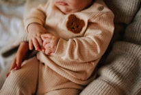 A close-up of a baby's torso dressed in a ribbed beige outfit adorned with a brown bear patch. The baby is gently holding onto an adult's hand, suggesting a tender moment. The adult's fingernails are painted red, adding a splash of color.