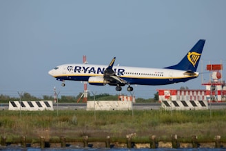 A commercial airplane in mid-landing approach on a runway, with airport equipment and signal lights in the background. The aircraft is branded with airline markings and is surrounded by grassy landscape.