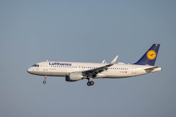 A Lufthansa passenger airplane is in flight, captured from a side view against a clear sky. The aircraft's landing gear is extended, and the Lufthansa logo is visible on the tail and fuselage.