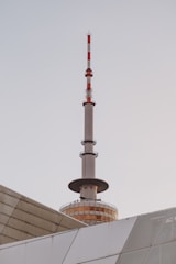 A tall communication tower with red and white stripes rises above a modern building with geometric patterns. The tower has a round, glass-enclosed observation deck near its base.