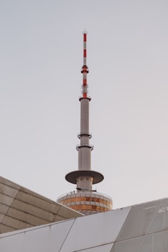 A tall communication tower with red and white stripes rises above a modern building with geometric patterns. The tower has a round, glass-enclosed observation deck near its base.