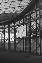 An interior view of a large, geometrically structured building with a glass ceiling and metal beams. Two signs are mounted on the metal framework, each displaying abstract figures with simple line designs. The floor is dark and textured, contrasting with the bright exterior visible through the windows.