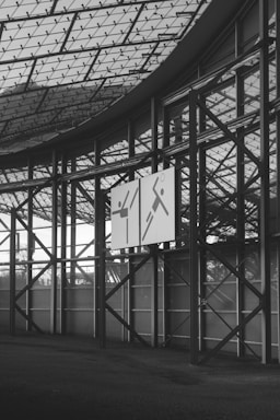 An interior view of a large, geometrically structured building with a glass ceiling and metal beams. Two signs are mounted on the metal framework, each displaying abstract figures with simple line designs. The floor is dark and textured, contrasting with the bright exterior visible through the windows.