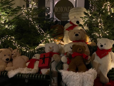 A group of plush teddy bears in various sizes are arranged on a bench surrounded by decorated Christmas trees with twinkling lights. Some bears are holding or sitting near red gift boxes, and most are wearing red scarves.