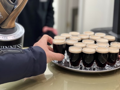 A hand reaching towards a tray filled with small glasses of dark stout, topped with a creamy head. The glasses are neatly arranged, and a beer tap featuring the brand is partially visible.