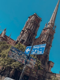 A tall, ornate building with intricate architectural details and pointed spires is seen in the background. In the foreground, a signpost covered in numerous colorful stickers points to Buenos Aires, with blue skies overhead.