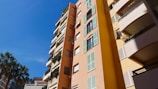 Tall residential buildings feature pastel-colored facades with green shutters and balconies adorned with plants. Palm trees stand nearby under a clear blue sky.