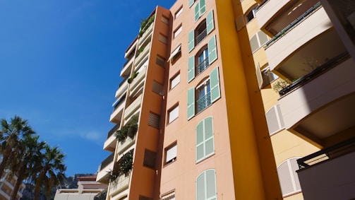 Tall residential buildings feature pastel-colored facades with green shutters and balconies adorned with plants. Palm trees stand nearby under a clear blue sky.