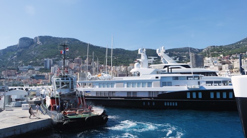 A large yacht is docked in a vibrant harbor, with a smaller boat moored nearby. The background features lush, green hills and tall buildings that make up a bustling coastal cityscape. Clear blue skies suggest pleasant weather, and a few people are visible on the dock and on the smaller boat, indicating activity and maritime operations.