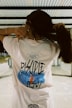 A person stands with their back to the camera, wearing a white shirt with the text 'Disciple of Jesus' on the back. The design includes a blue and orange graphic element. The person is holding their long hair up with both hands. The background features a white and gray industrial-looking structure with metal beams.