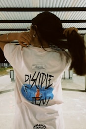 A person stands with their back to the camera, wearing a white shirt with the text 'Disciple of Jesus' on the back. The design includes a blue and orange graphic element. The person is holding their long hair up with both hands. The background features a white and gray industrial-looking structure with metal beams.