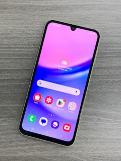 A smartphone with a colorful display featuring various app icons such as Store, Gallery, Google Play, and Google, is lying on a wooden surface. The screen has a gradient background of pink and purple shades.