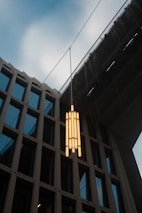 An outdoor view featuring a contemporary pendant light hanging between a modern building and an overpass. The light fixture comprises multiple cylindrical bulbs arranged vertically. The building has a grid of large windows framed by textured concrete. The sky is slightly overcast, adding a subtle atmospheric effect.