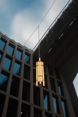 An outdoor view featuring a contemporary pendant light hanging between a modern building and an overpass. The light fixture comprises multiple cylindrical bulbs arranged vertically. The building has a grid of large windows framed by textured concrete. The sky is slightly overcast, adding a subtle atmospheric effect.