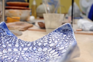A detailed close-up of a ceramic bowl with an intricate blue lace pattern against a blurred background of a pottery studio. The craftsmanship and texture of the bowl are prominent.