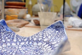 A detailed close-up of a ceramic bowl with an intricate blue lace pattern against a blurred background of a pottery studio. The craftsmanship and texture of the bowl are prominent.