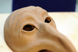 A close-up view of a clay mask with a long, beak-like nose and hollow eyes, resting on a light wooden surface. The mask appears to be in the style reminiscent of a historical plague doctor mask.