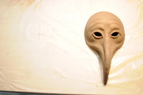 A clay mask with exaggerated facial features, including a long, pointed nose and hollow eyes, placed on a light beige surface. The mask has a smooth, natural texture that resembles a bird's beak.