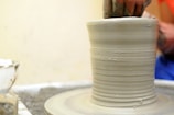 A person is shaping a tall, cylindrical clay object on a pottery wheel with their hands. The image captures the textured surface of the clay as it spins, with a focus on the craftsmanship involved in pottery making.