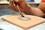A hand skillfully holds a paintbrush, applying paint to a rectangular surface. The focus is on the hand and brush, creating a circular pattern on a clay-colored surface.