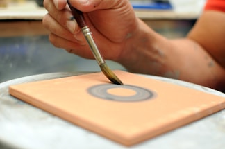 A hand skillfully holds a paintbrush, applying paint to a rectangular surface. The focus is on the hand and brush, creating a circular pattern on a clay-colored surface.