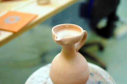A handcrafted clay jug with a curved handle and unfinished surface rests on a pottery wheel. The background is slightly blurred, with hints of a workspace including an orange notebook.