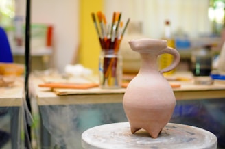 On a pottery wheel, a clay vessel with a spout and handle is prominently displayed. The background shows brushes in a jar and blurred objects, giving the appearance of a creative studio environment.