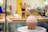 On a pottery wheel, a clay vessel with a spout and handle is prominently displayed. The background shows brushes in a jar and blurred objects, giving the appearance of a creative studio environment.