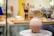 On a pottery wheel, a clay vessel with a spout and handle is prominently displayed. The background shows brushes in a jar and blurred objects, giving the appearance of a creative studio environment.