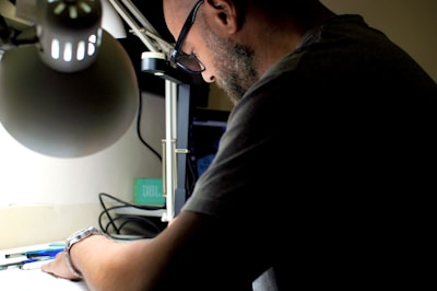 A person with a beard and glasses is focused on an activity at a desk. A desk lamp illuminates the workspace, casting light on the person and a collection of pens and papers. The background is dimly lit, suggesting a calm and concentrated environment.