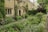 A charming stone house surrounded by a lush, well-maintained garden. The exterior features large windows, and the garden is filled with a variety of flowering plants and bushes. Ivy climbs up the house walls, adding to the cottage-like aesthetic. A pathway runs alongside the garden, lined with dense greenery.